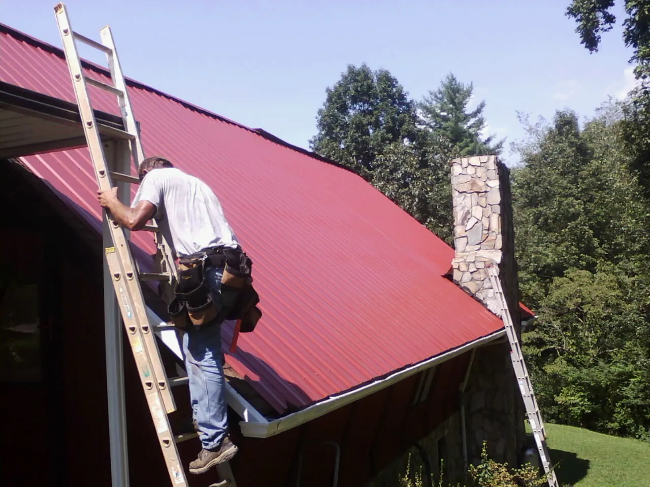 Roofer on a ladder beside a red metal roof and stone chimney — Soffit & Fascia Repair by Roofing Brothers in Indian Orchard